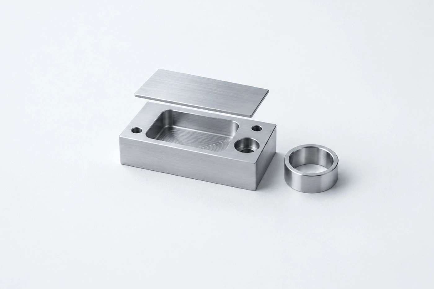 Illustrative machined aluminium block with a lifted slice, open pocket and bore, beside a turned ring.